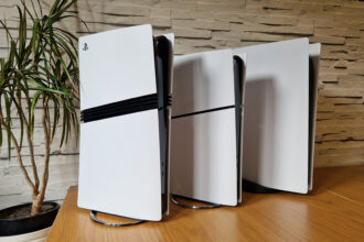 The PS5 Pro console next to a PS5 Slim and launch PS5 on a wooden desk in front of a white brick background and next to a potted plant