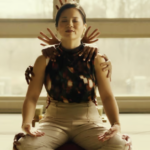 A still from the Hulu movie Control Freak of Kelly Marie Tran sitting cross-legged as hands appear behind her