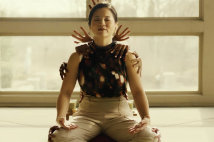 A still from the Hulu movie Control Freak of Kelly Marie Tran sitting cross-legged as hands appear behind her