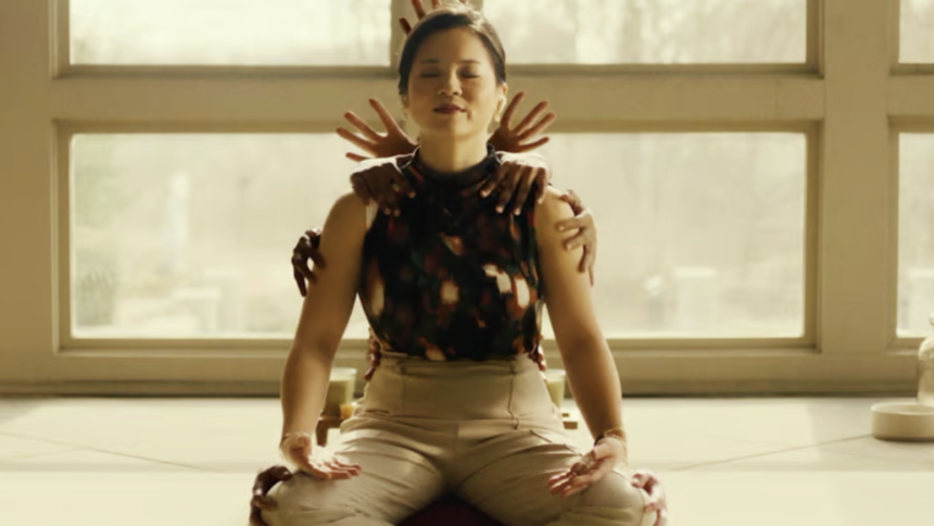 A still from the Hulu movie Control Freak of Kelly Marie Tran sitting cross-legged as hands appear behind her