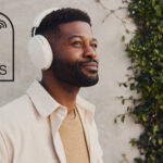 A man wearing a white pair of Sonos Ace headphones, standing in front of a wall. The TechRadar logo and &quot;Big Savings&quot; text are in black, in the top right.