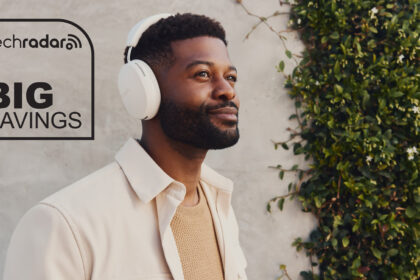 A man wearing a white pair of Sonos Ace headphones, standing in front of a wall. The TechRadar logo and &quot;Big Savings&quot; text are in black, in the top right.