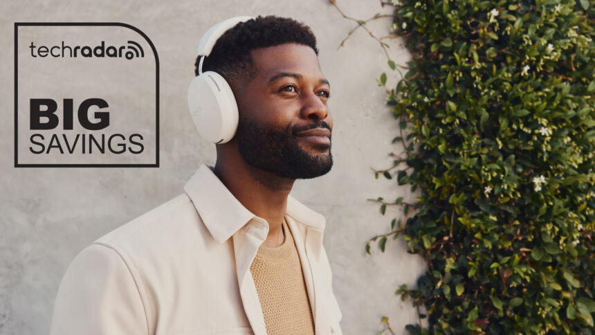 A man wearing a white pair of Sonos Ace headphones, standing in front of a wall. The TechRadar logo and &quot;Big Savings&quot; text are in black, in the top right.