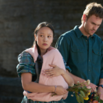 A still from the Netflix series The Survivors showing a man and a woman on a beach with a cliff behind them. The woman is carrying a baby in a pink blanket, although the baby is not visible.