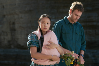 A still from the Netflix series The Survivors showing a man and a woman on a beach with a cliff behind them. The woman is carrying a baby in a pink blanket, although the baby is not visible.