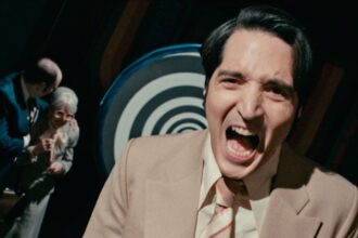 David Dastmalchian laughs maniacally in front of a hypnotic background in Late Night with the Devil