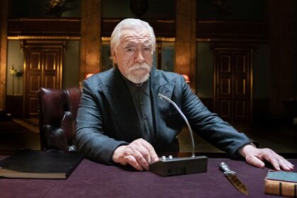 Brian Cox sits at a desk pressing a button on an intercom