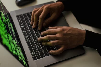 A hacker typing on a MacBook laptop with code on the screen.