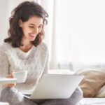 Happy woman sitting on a bed with a coffee and a Windows 10 laptop