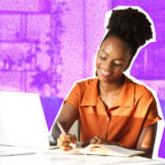 A woman sitting at a desk, writing in a notepad. A laptop is also on the table.