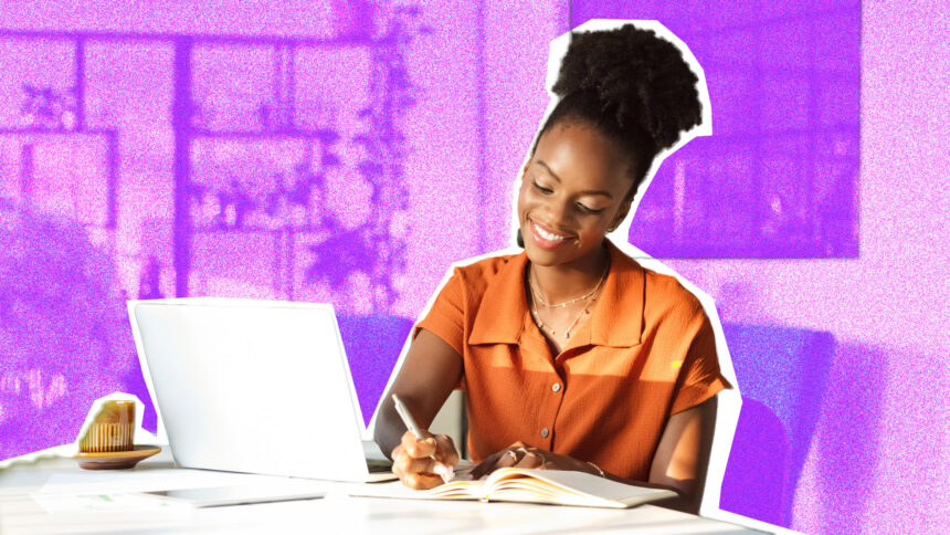 A woman sitting at a desk, writing in a notepad. A laptop is also on the table.