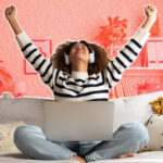 A young woman sitting on a sofa, wearing headphones and throwing her arms excitedly into the air.