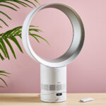 The Dyson Cool CF1 is facing slightly to the right on a table top in front of a leafy green plant and a pink wall. The remote control is on the table to the right of the fan.