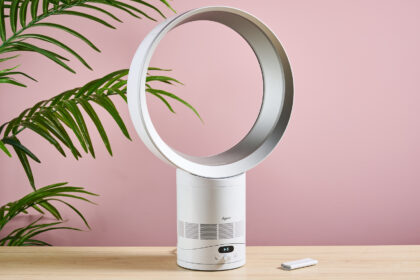 The Dyson Cool CF1 is facing slightly to the right on a table top in front of a leafy green plant and a pink wall. The remote control is on the table to the right of the fan.