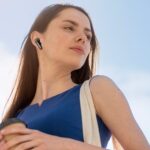 A young woman in a blue shift dress with dark brown hair is listening to a pair of EarFun Air Pro 4+ earbuds against a blue and slightly cloudy sky