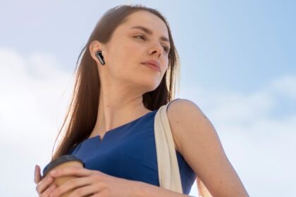 A young woman in a blue shift dress with dark brown hair is listening to a pair of EarFun Air Pro 4+ earbuds against a blue and slightly cloudy sky