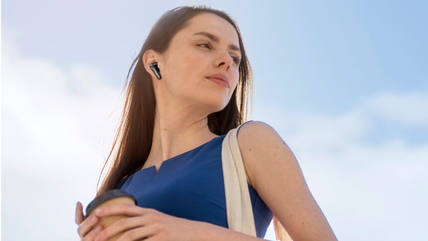 A young woman in a blue shift dress with dark brown hair is listening to a pair of EarFun Air Pro 4+ earbuds against a blue and slightly cloudy sky