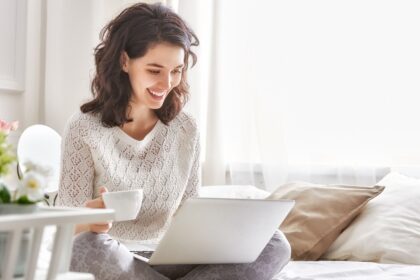 Happy woman sitting on a bed with a coffee and a laptop