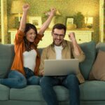 Young couple looking excited whilst sitting on sofa with a laptop.