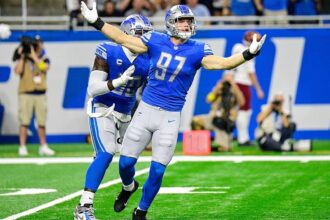 Aidan Hutchinson celebrates with arms wide open on the football field during a Detroit Lions game.