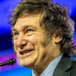 Javier Milei delivering a passionate speech on stage, gesturing emphatically to the audience under bright stage lights.