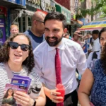 Zohran Mamdani taking selfies with supporters at a campaign event, smiling and engaging with the crowd.