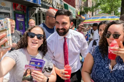 Zohran Mamdani taking selfies with supporters at a campaign event, smiling and engaging with the crowd.