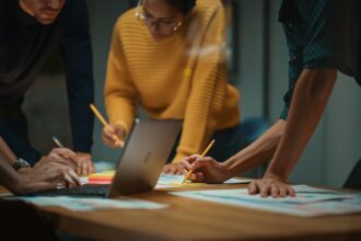 close up of diverse multiethnic team having conversation in meeting room in a creative office. colleagues lean on a conference table, look at laptop computer and make notes with pencils on notebooks.