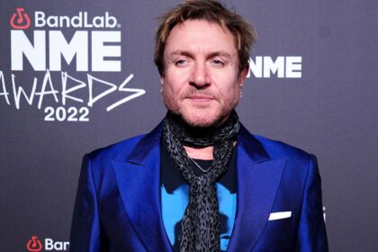 Simon Le Bon attends the NME Awards 2022 wearing a blue satin blazer and patterned scarf, posing on the red carpet.