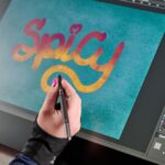 A woman's hand is holding a slim stylus over the drawing display, she is putting the finishing touches on some bright brush lettering that says &quot;Spicy&quot;.