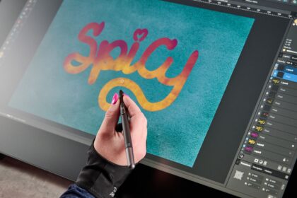 A woman's hand is holding a slim stylus over the drawing display, she is putting the finishing touches on some bright brush lettering that says &quot;Spicy&quot;.