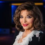 Dame Joan Collins smiling in an elegant black jacket with a white ruffled blouse, wearing a diamond brooch, photographed under soft blue lighting at a London event.