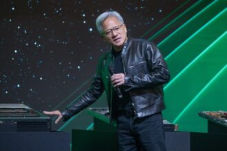 Jensen Huang, CEO of Nvidia, presenting on stage in a black leather jacket, holding a microphone and gesturing toward a GPU system.