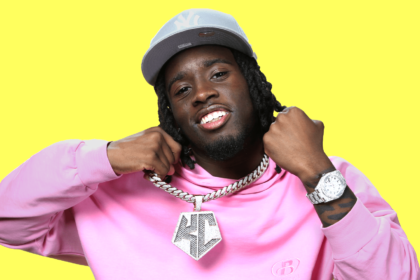 Kai Cenat posing confidently with his diamond chain and luxury watch against a vibrant yellow background.