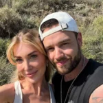 Liam Payne and Kate Cassidy, smiling and laughing as they take a selfie together on a sunny hiking trail surrounded by lush green hills and wildflowers.