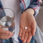 Person holding a glass of water and a single supplement pill in their hand.