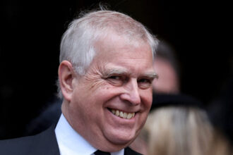 Prince Andrew smiling while attending a recent royal funeral, dressed in formal attire.