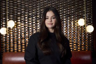 Selena Gomez poses for a portrait wearing a black top, standing in front of a patterned metallic backdrop with hanging lights.