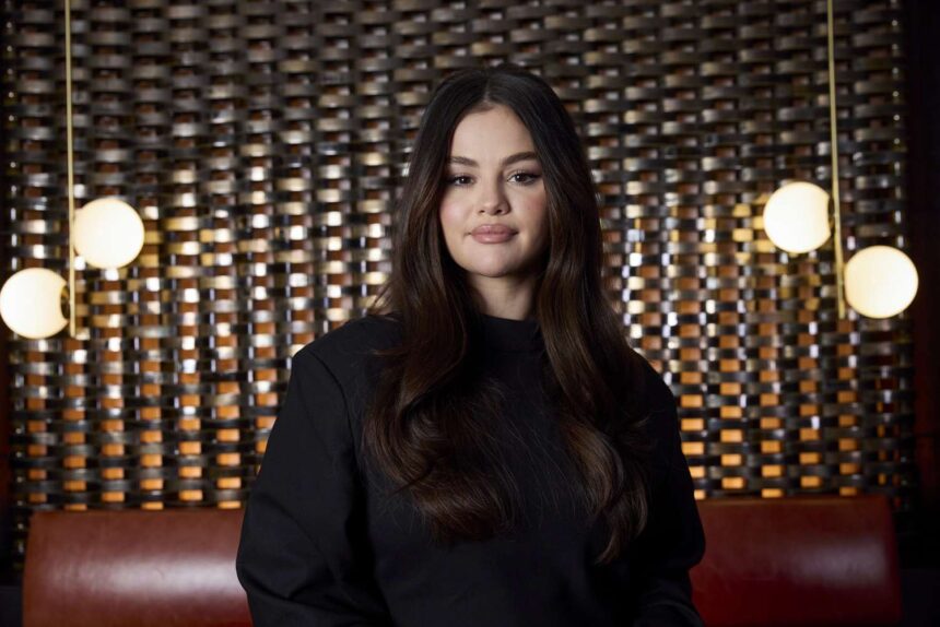 Selena Gomez poses for a portrait wearing a black top, standing in front of a patterned metallic backdrop with hanging lights.