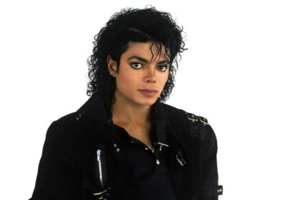 Michael Jackson wearing his iconic black leather jacket, standing against a white background, striking a classic pose.