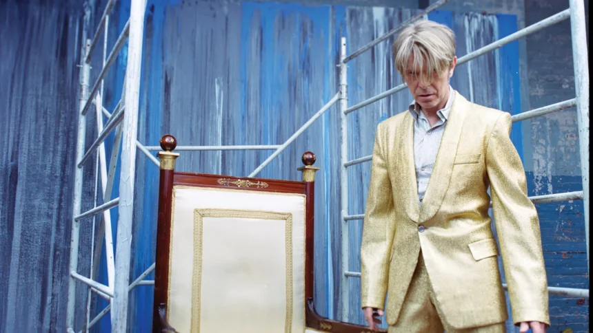 David Bowie wearing a gold suit beside a decorative chair on a blue-toned set, evoking his late-era artistic src and visual experimentation.
