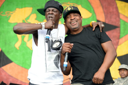 Flavor Flav and Chuck D of Public Enemy performing on stage, standing close together with microphones, smiling in front of a colorful backdrop.