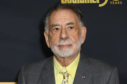 Francis Ford Coppola attending a film industry event, wearing a gray suit and yellow shirt, photographed against a black backdrop.