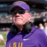 Brian Kelly, LSU Tigers head coach, in purple and gold team gear, wearing headphones and sunglasses during a high-energy practice session.