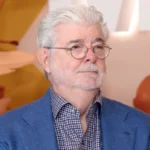 George Lucas smiling warmly on the red carpet at a film premiere, dressed in a dark suit and patterned tie.
