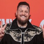 Jelly Roll flashing a peace sign while posing on the red carpet, wearing a black suit and sunglasses.