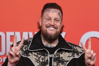 Jelly Roll flashing a peace sign while posing on the red carpet, wearing a black suit and sunglasses.