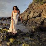 Kim Kardashian standing on a rocky beach in a form-fitting white dress, gazing out at the sea, radiating calm and confidence on bar exam results day.