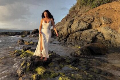 Kim Kardashian standing on a rocky beach in a form-fitting white dress, gazing out at the sea, radiating calm and confidence on bar exam results day.