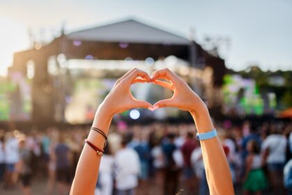 concertgoer forms heart shape with hands at beachside music festival, crowd enjoys live performance in sunset, love symbol gesture at summer event, youth celebration of art, outdoor entertainment.